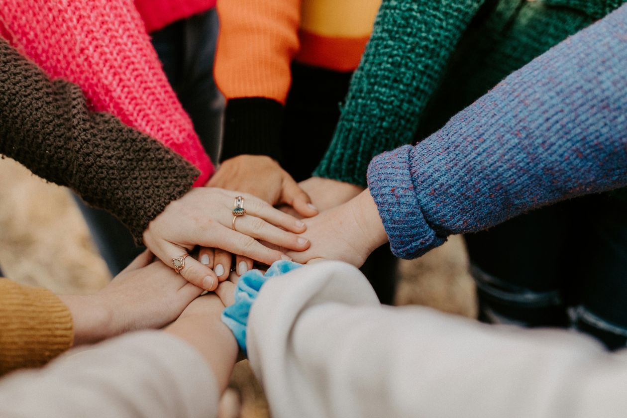 multiple hands one on top of another in a circle with sweater sleeves in green, pink, orange, blue, white, brown and gray